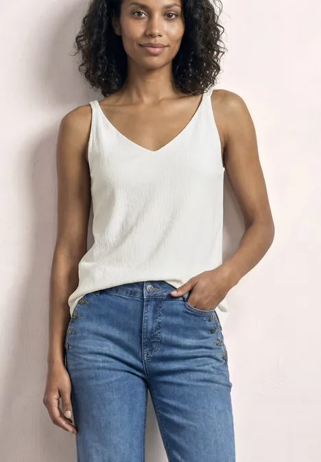 Frau mit lockigen Haaren in weißem Top und blauen Jeans, steht entspannt.